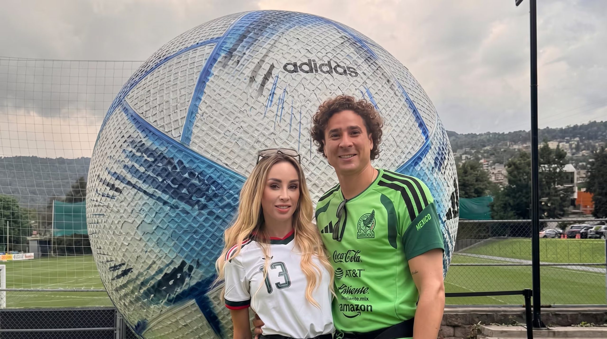 Guillermo Ochoa y Karla Mora sonríen mientras posan frente a un balón de fútbol gigante Adidas en un campo deportivo con malla. (Imagen Ilustrativa Infobae)