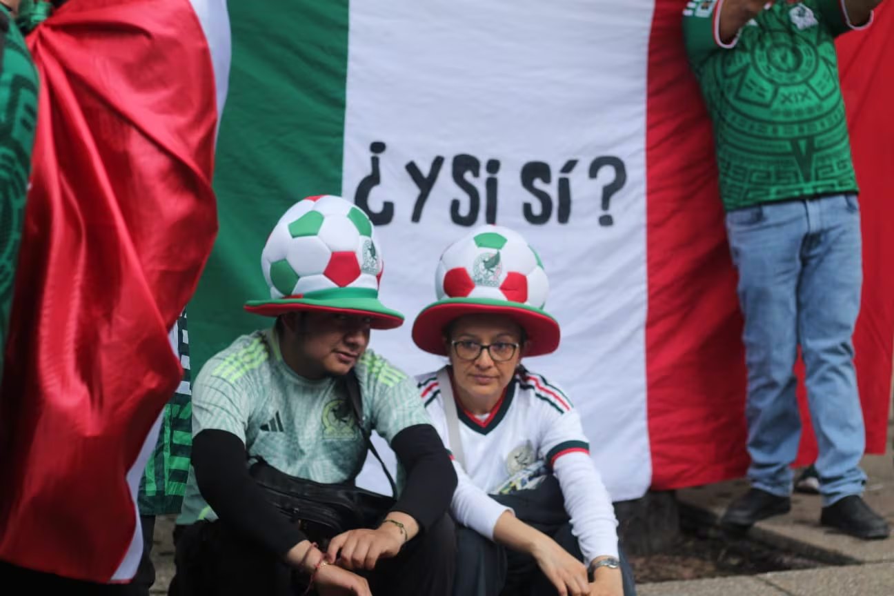Dos aficionados con camisetas y sombreros de fútbol con los colores de México se muestran frente a una bandera nacional que exhibe la frase 