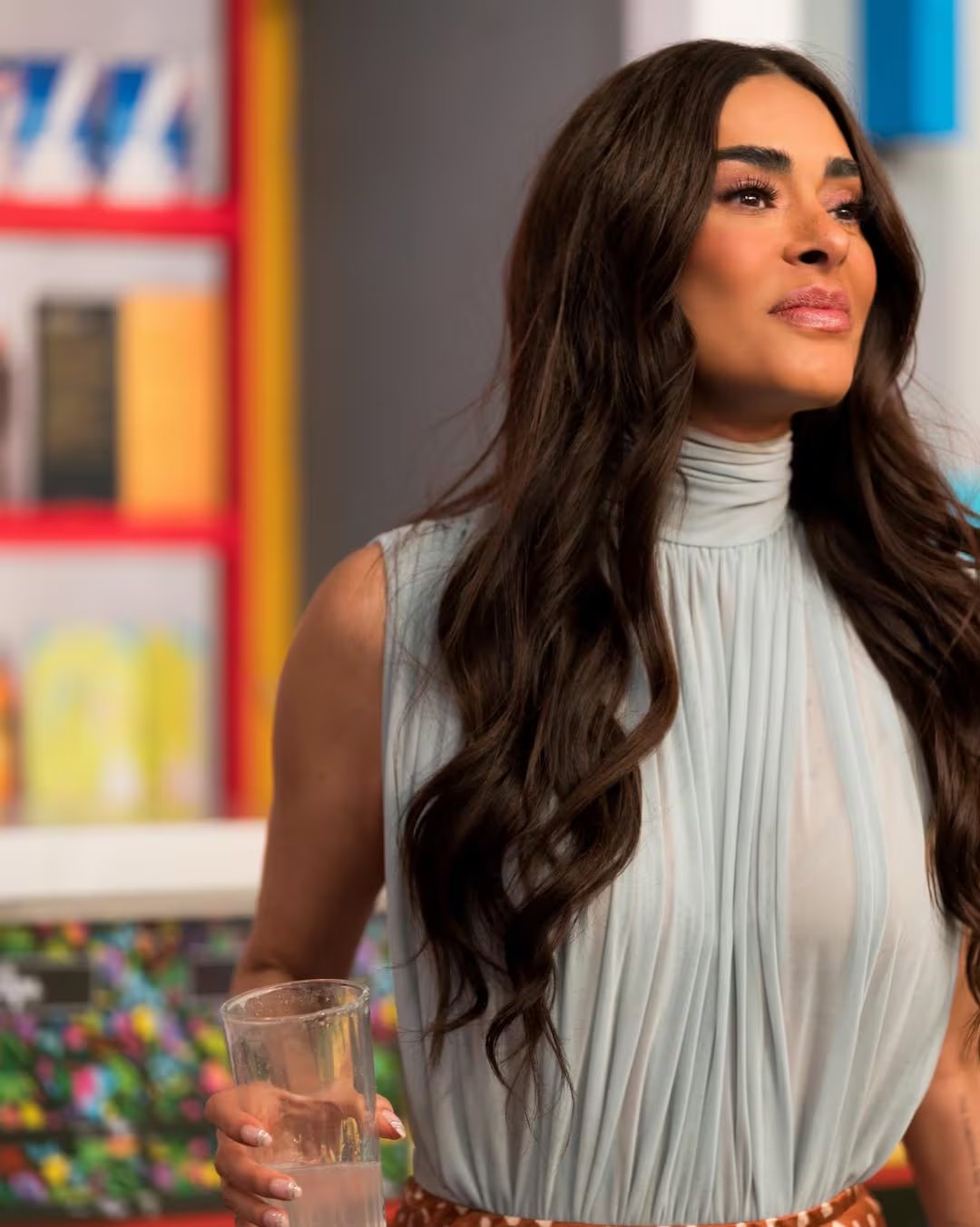 Una mujer con cabello oscuro y blusa celeste sostiene un vaso de agua, observando pensativa su entorno. (Galilea Montijo/Instagram)