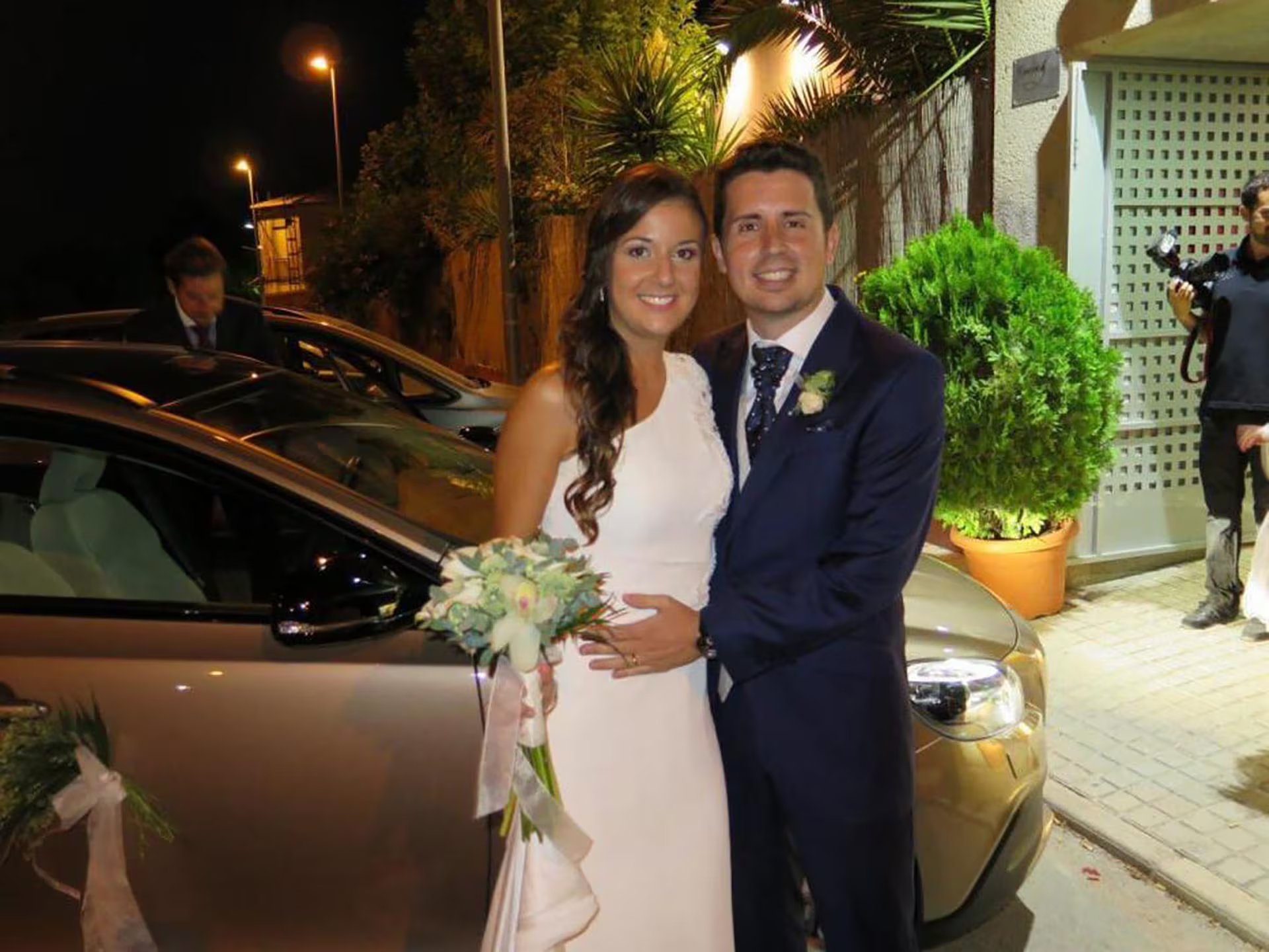 Maje Moreno Cantó y su pareja, vestidos con traje nupcial, posan sonrientes en el exterior junto a un vehículo decorado para la celebración.