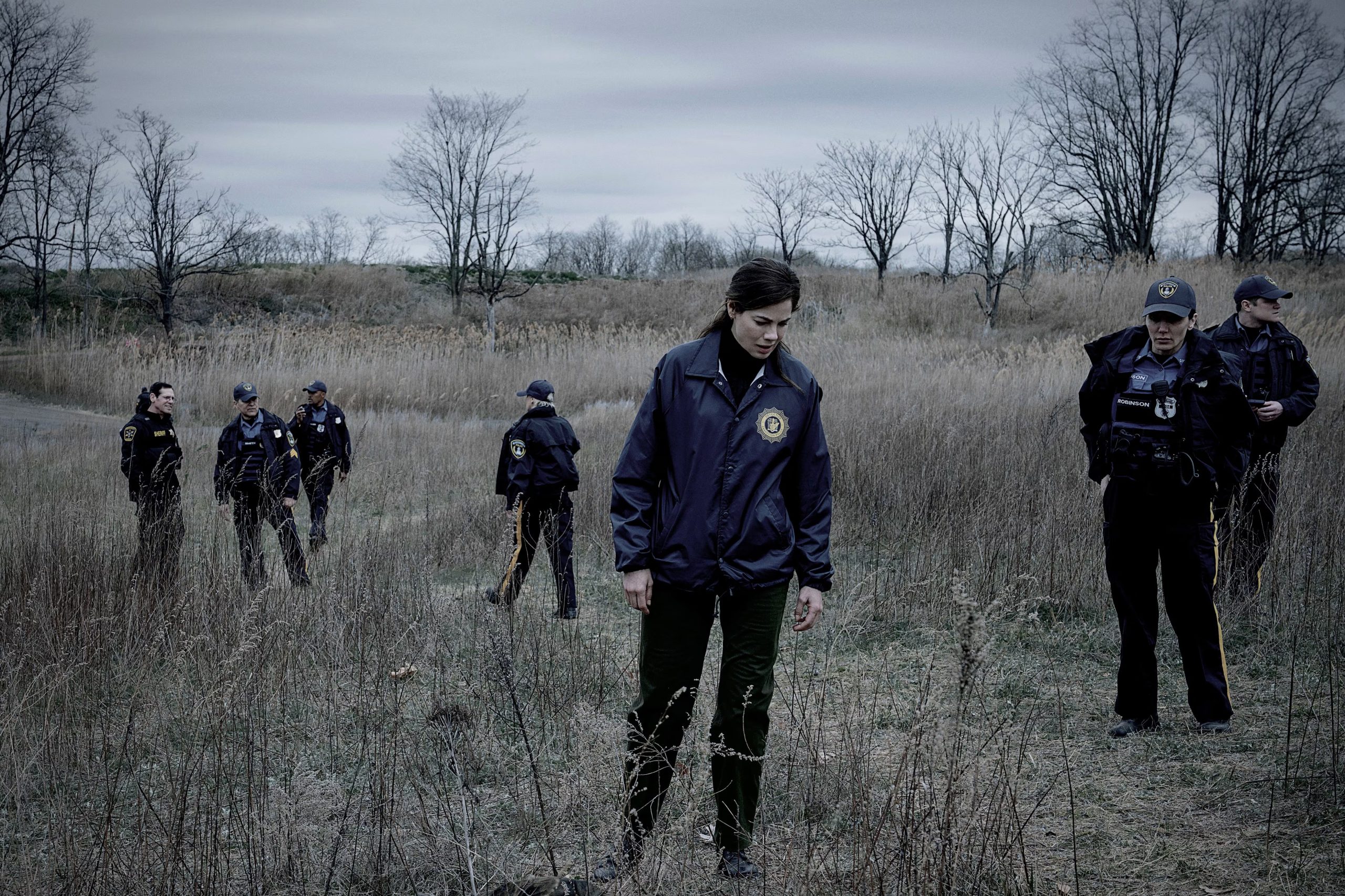 La actriz Michelle Monaghan, interpretando a una oficial de policía, avanza por un campo de hierba seca junto a otros agentes policiales bajo un cielo nublado. (Netflix)