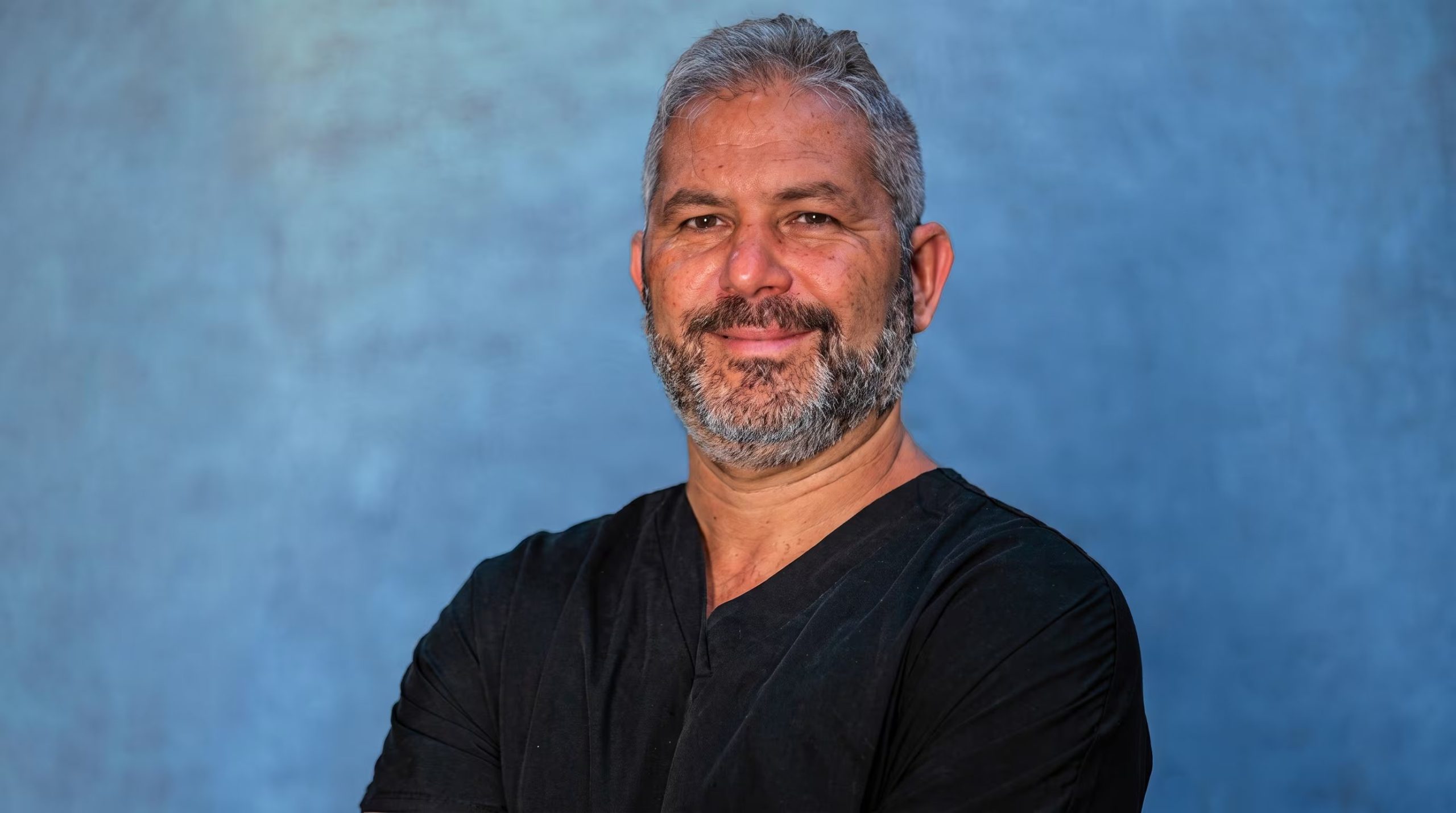 Un hombre con cabello canoso y barba, vestido con una camisa oscura de cuello en V, sonríe y mira directamente a la cámara sobre un fondo azul texturizado. (Imagen Ilustrativa Infobae)