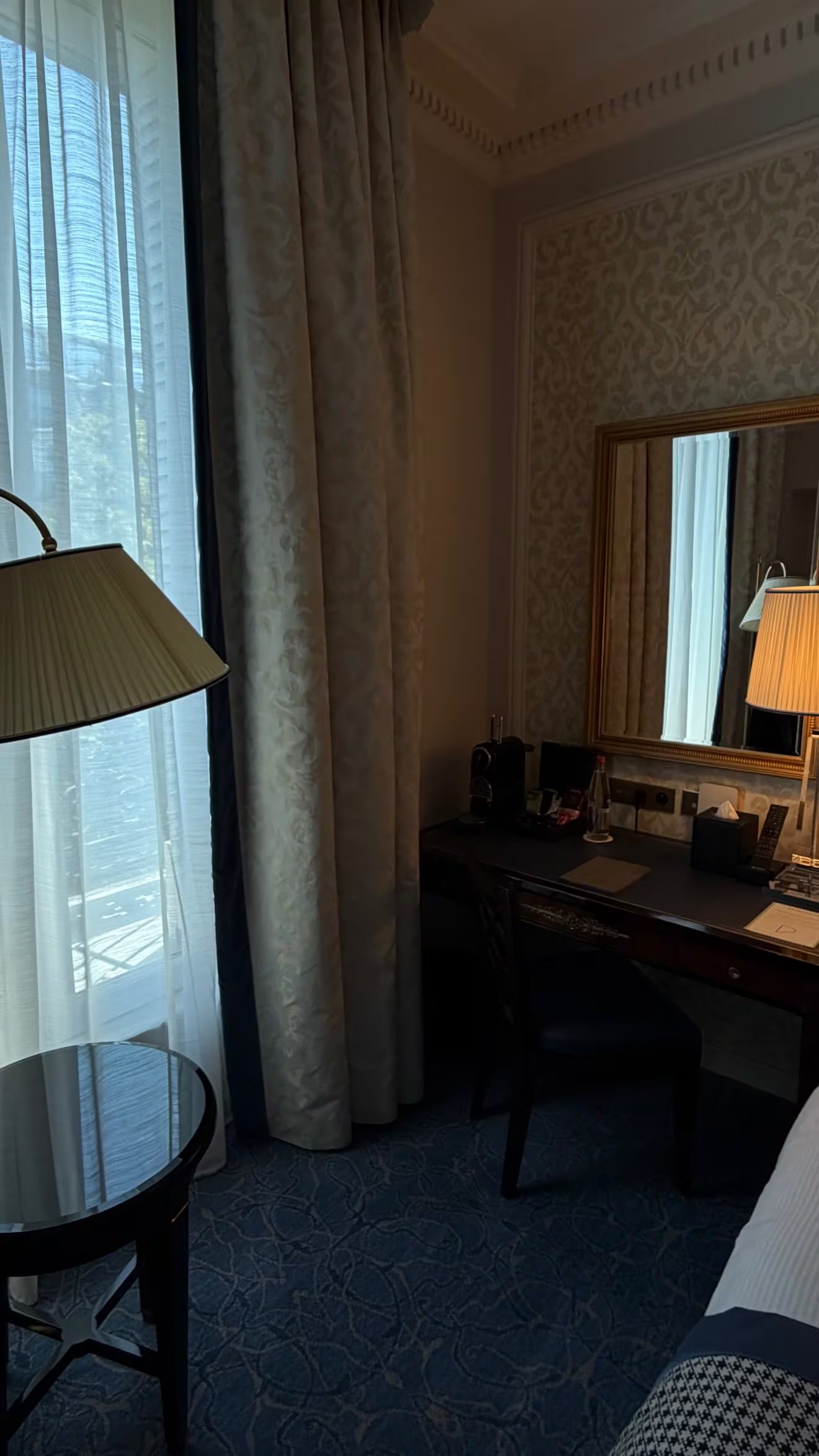 Una habitación de hotel en París muestra un escritorio con silla, una lámpara de mesa y un espejo de marco dorado junto a una ventana con cortinas.