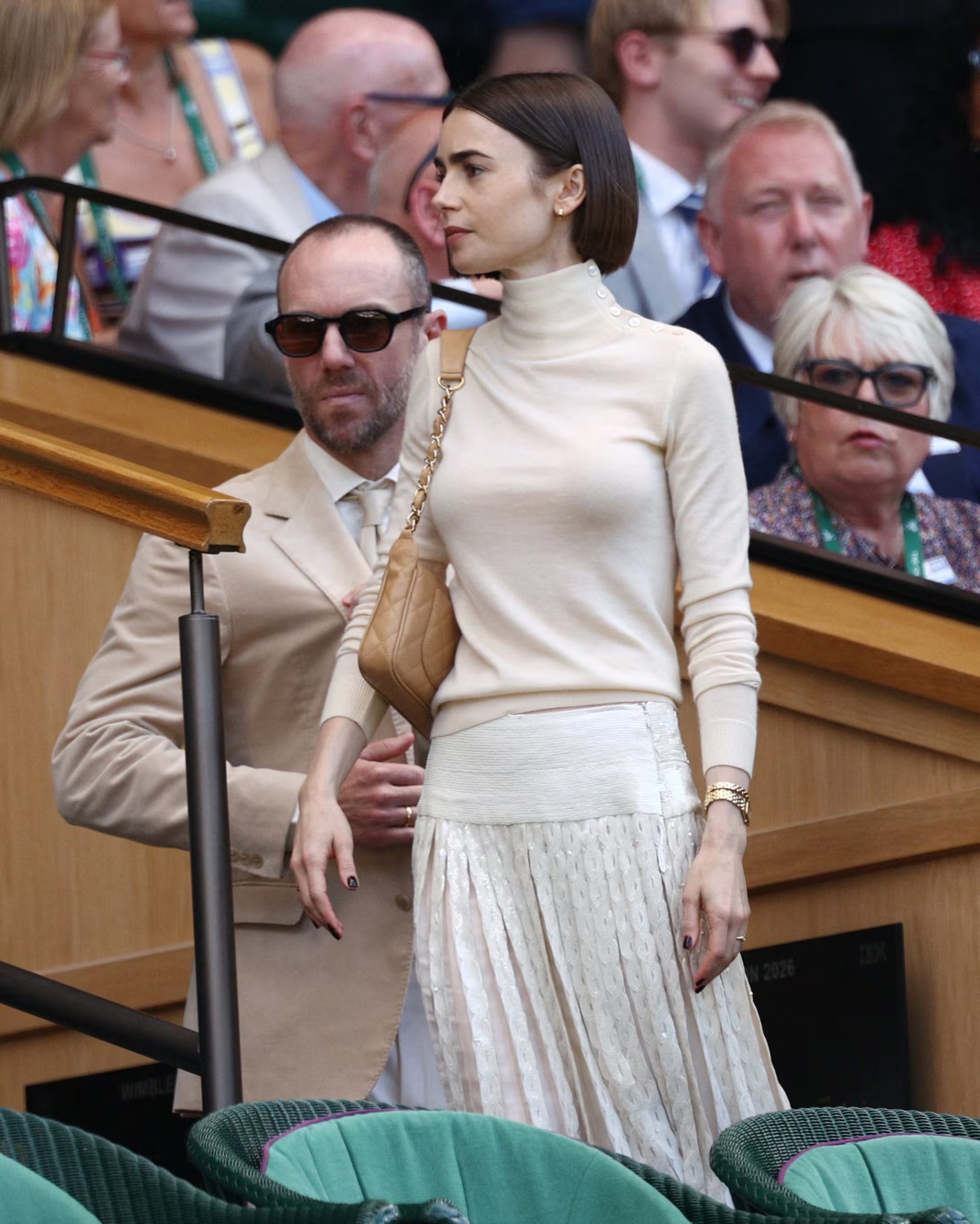 Lily Collins combina crema y beige con falda plisada, accesorios dorados, uñas oscuras y maquillaje discreto