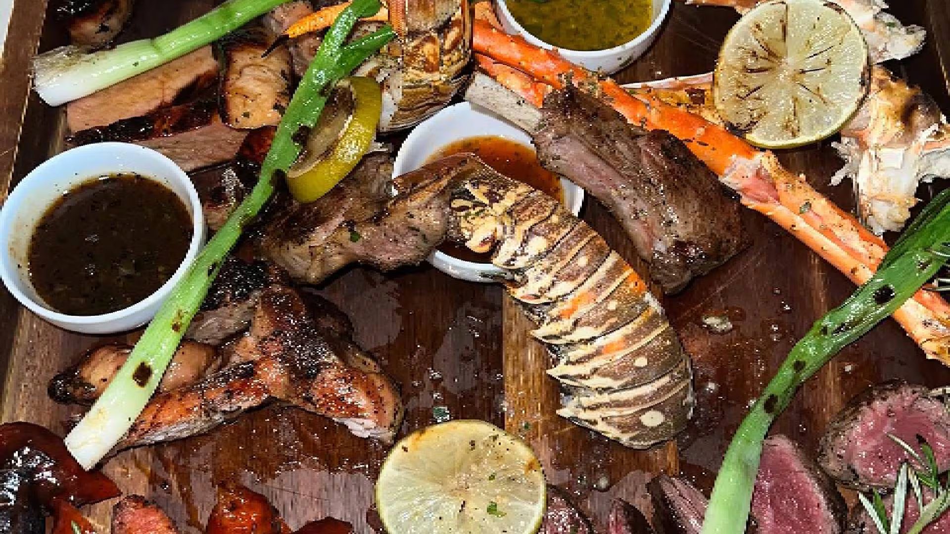 Un variado plato principal presenta langosta, patas de cangrejo y cortes de carne roja a la parrilla, acompañados de limón, cebolla verde y varias salsas ( C.L.A.S.S. Soiree Steakhouse)