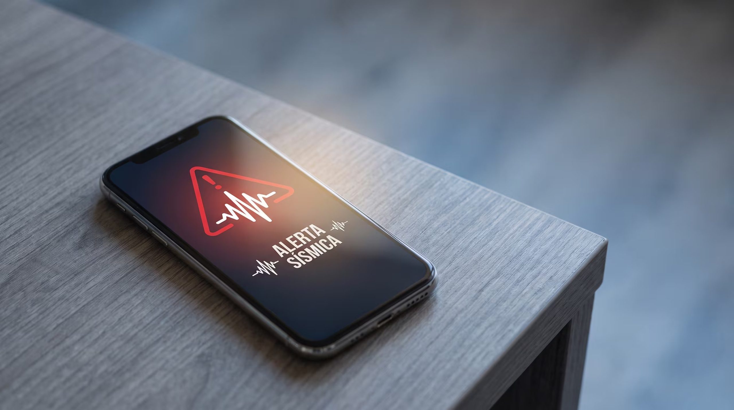 Un teléfono móvil moderno sobre una mesa exhibe una alerta sísmica con un símbolo de onda y un triángulo de advertencia rojo en su pantalla. (Imagen Ilustrativa Infobae)