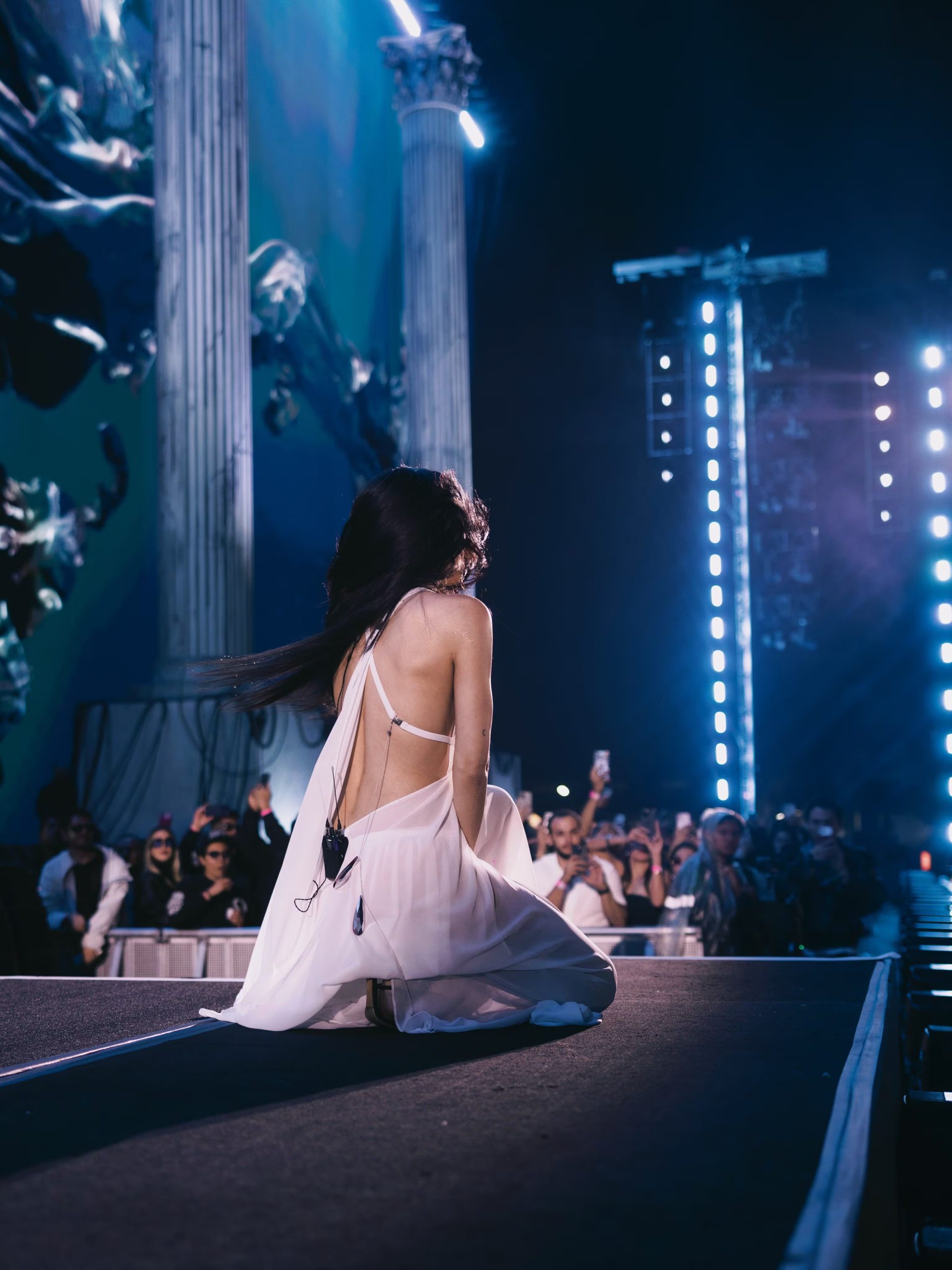 Una artista con un vestido blanco y la espalda al aire permanece arrodillada en la pasarela de un escenario ante el público de un concierto. (Kenia Os/Instagram)