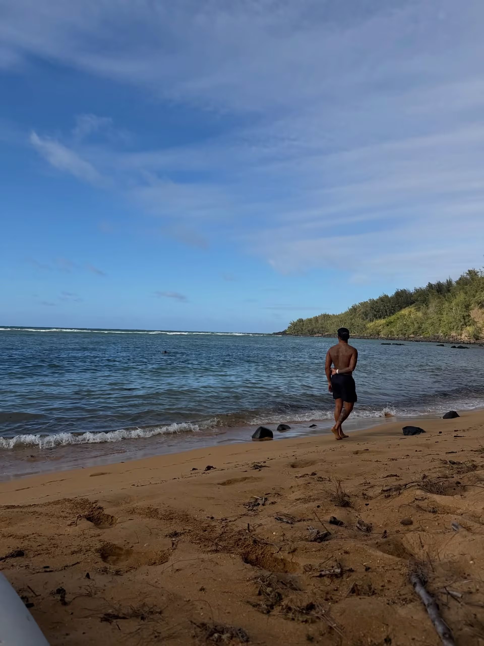 Lewis Hamilton camina por la orilla de una playa de arena dorada y aguas tranquilas bajo un cielo parcialmente nublado