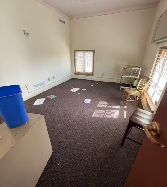 Una estancia casi vacía conserva un contenedor azul, sillas de madera y hojas de papel esparcidas sobre una alfombra oscura. (Tim Walters)