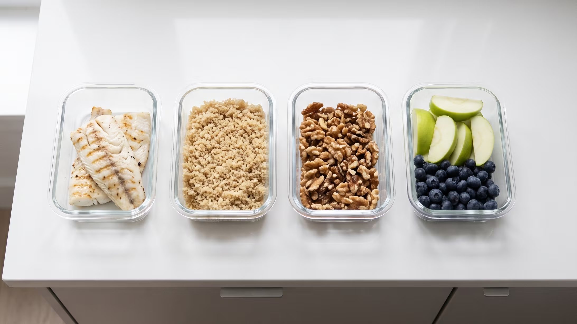 Cuatro tápers transparentes sobre una encimera blanca muestran la organización de comidas con porciones de pescado grillado, quinoa, nueces y una mezcla de manzana verde y arándanos. (Imagen Ilustrativa Infobae)