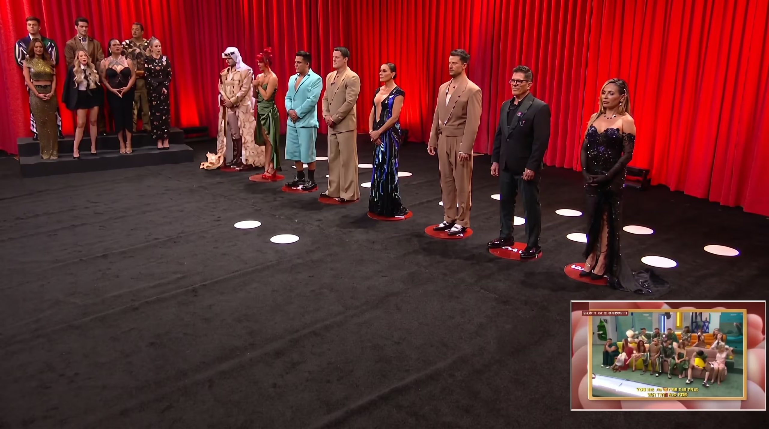 Un grupo de concursantes vestidos de gala permanece de pie sobre un escenario de televisión con fondo de cortinas rojas. (Imagen Ilustrativa Infobae)