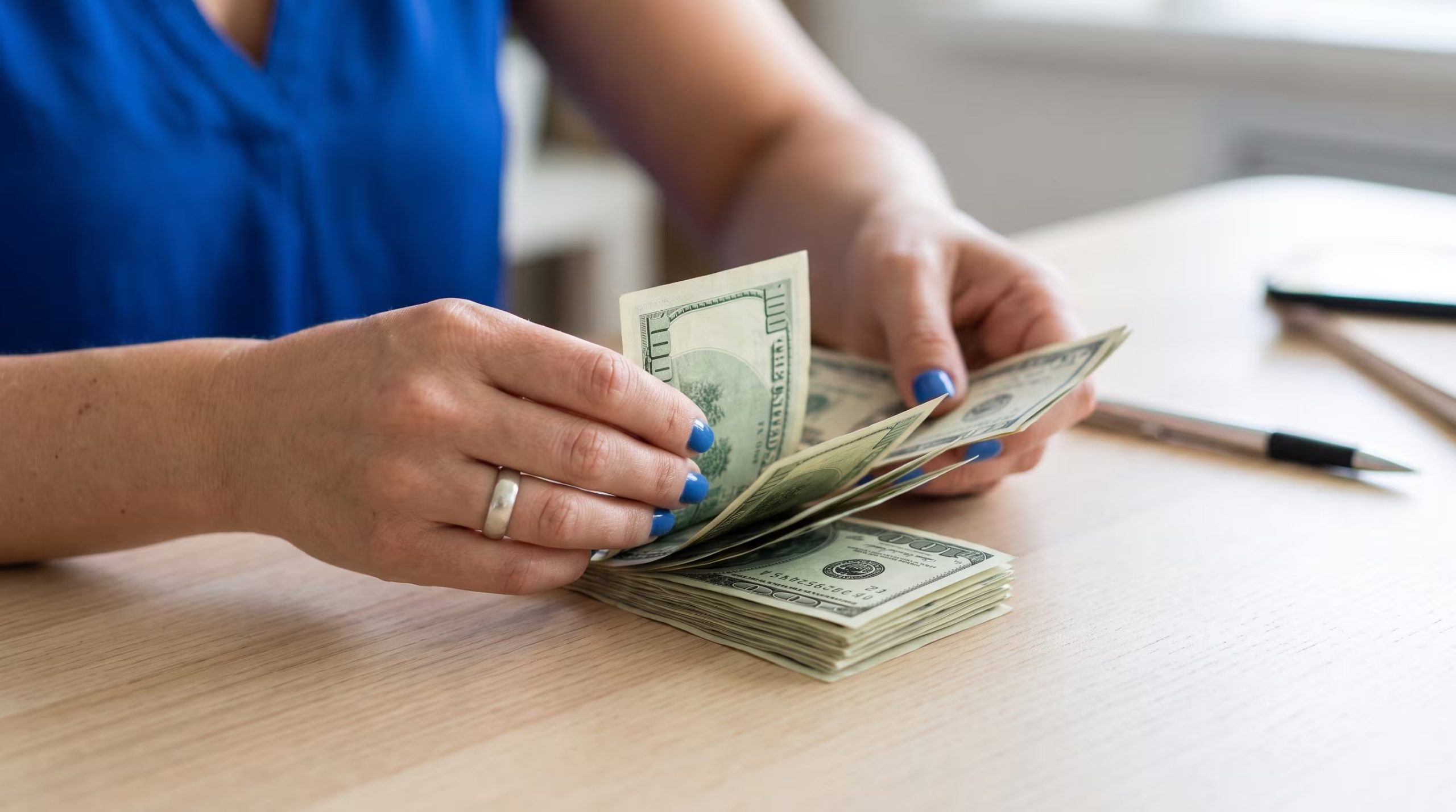 Una persona con esmalte azul en las uñas manipula billetes de cien dólares, ordenados en un fajo sobre una superficie de madera clara. (Imagen Ilustrativa Infobae)