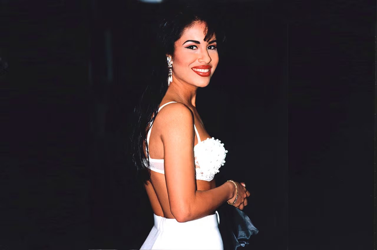 Selena Quintanilla, la icónica cantante de Tex-Mex, posa sonriente luciendo un top blanco y accesorios brillantes.