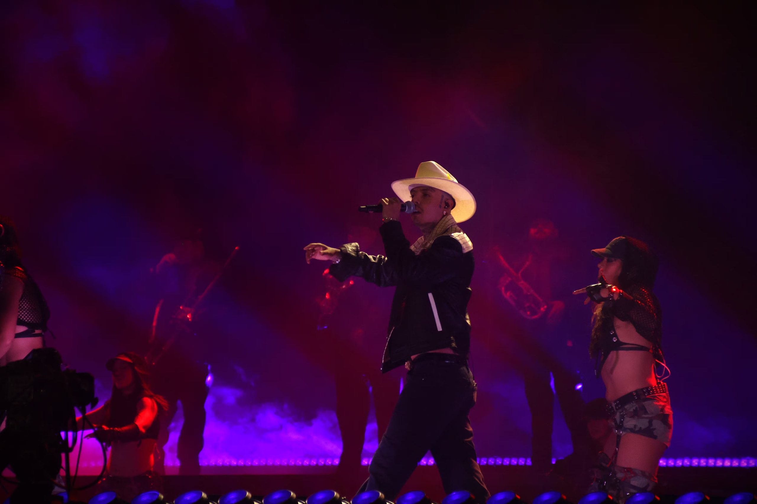 Un cantante con sombrero realiza una interpretación musical acompañado por bailarines y músicos bajo reflectores de color morado. (Selene Miranda/Infobae México)