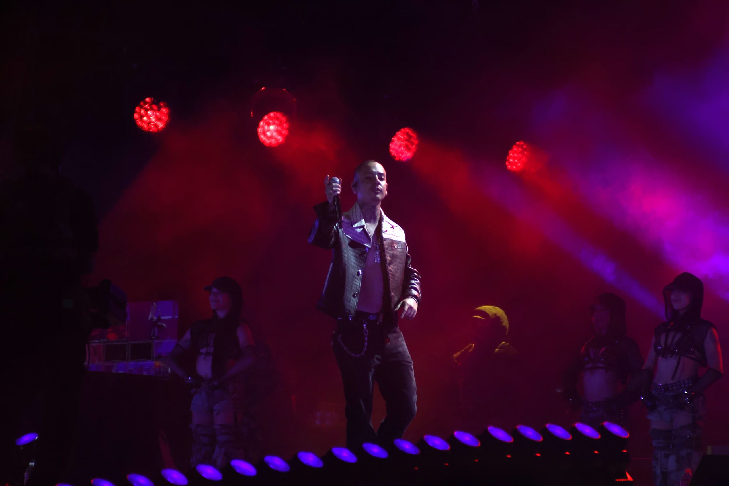 Un cantante sostiene un micrófono sobre un escenario iluminado por luces rojas durante un concierto en vivo. (Selene Miranda/Infobae México)