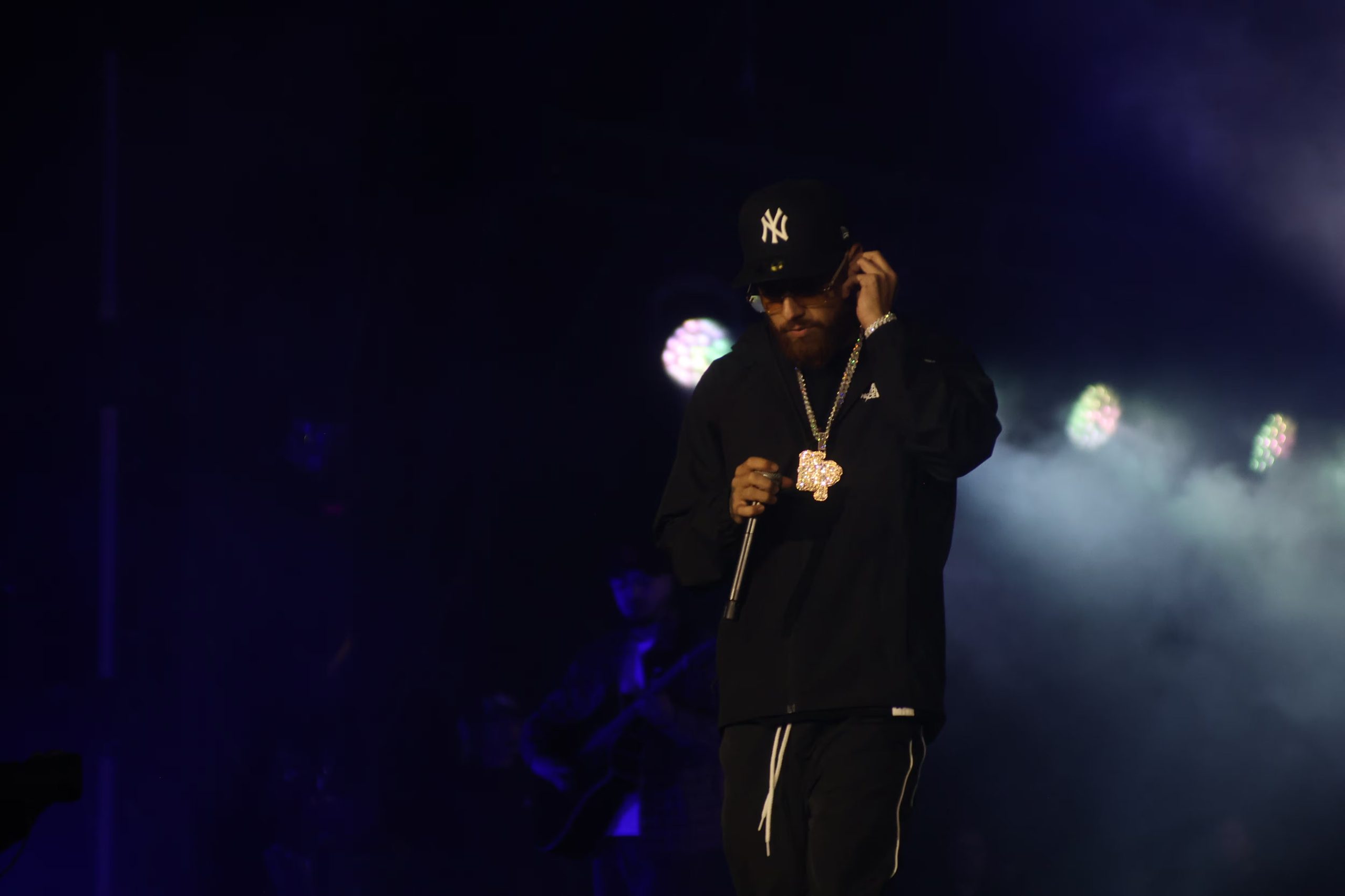 Un cantante con gorra negra, gafas y un collar grande sostiene un micrófono durante una presentación sobre un escenario iluminado. (Selene Miranda/Infobae México)
