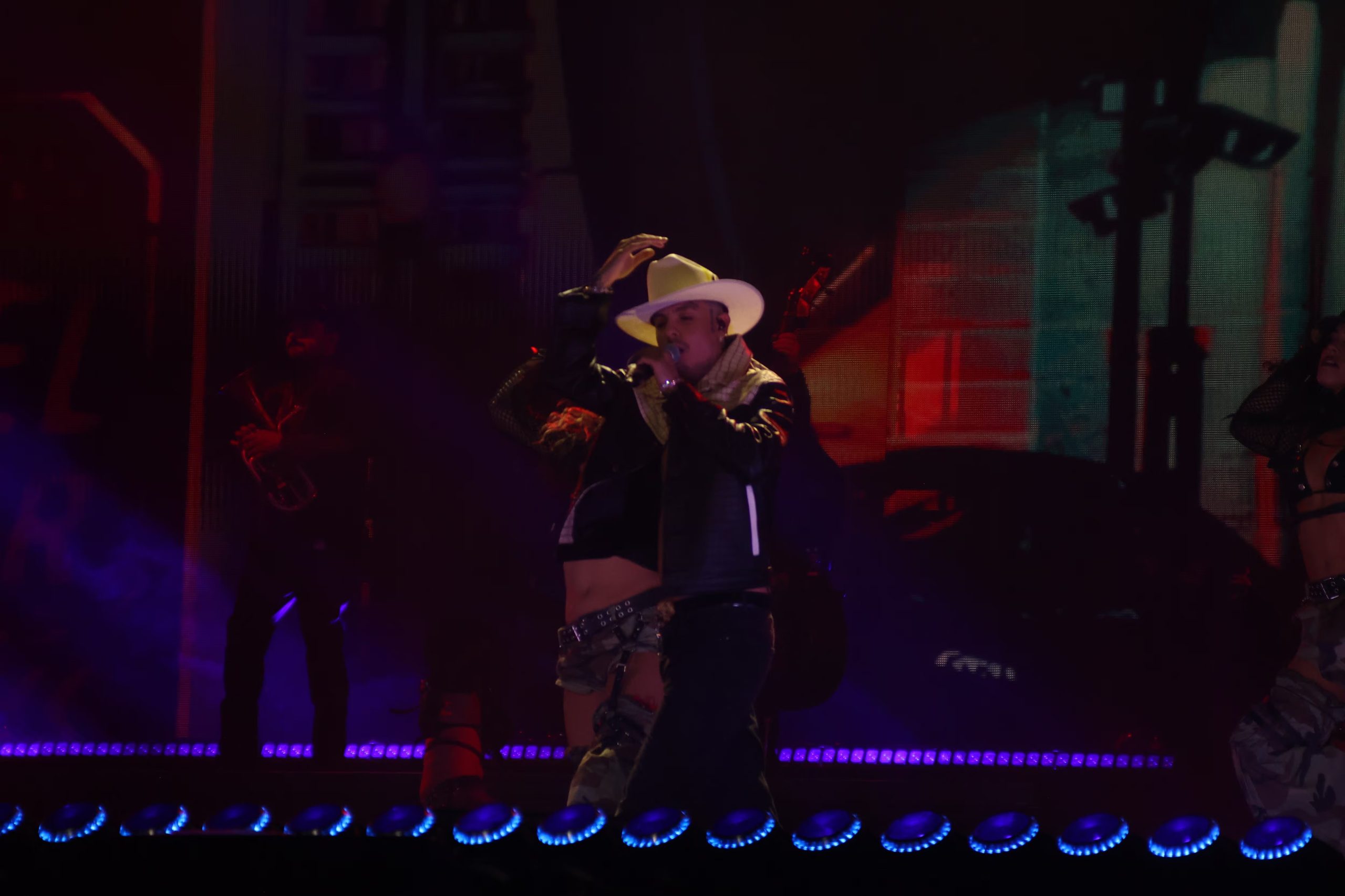 Un cantante con sombrero blanco y chaqueta oscura interpreta una canción durante una presentación en un escenario iluminado. (Selene Miranda/Infobae México)