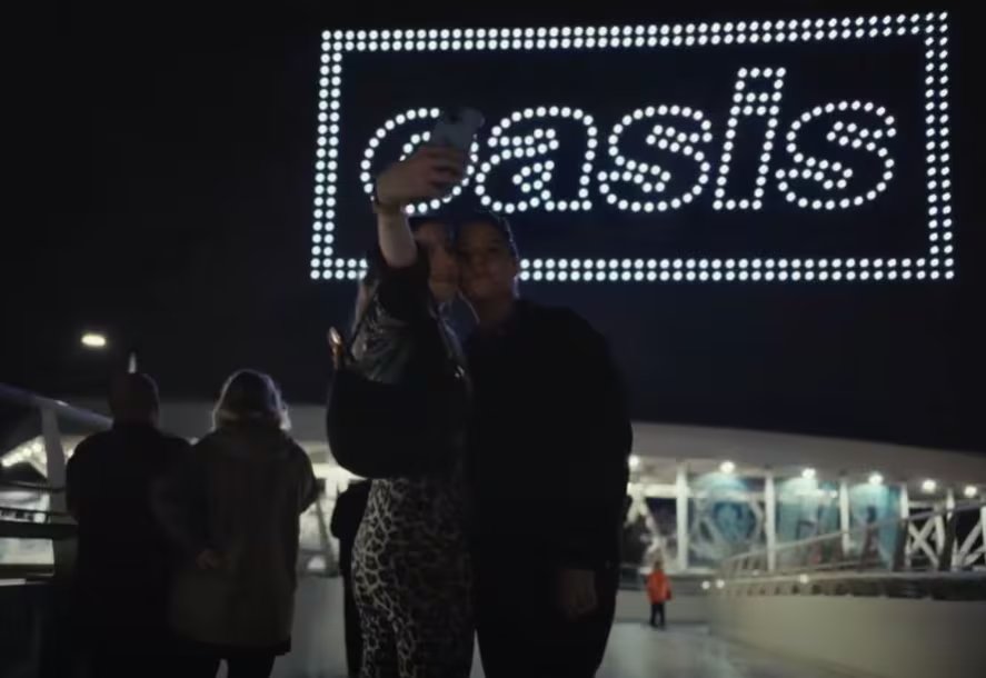 Dos asistentes se toman una fotografía nocturna mientras un espectáculo de drones proyecta el logotipo del grupo Oasis en el cielo de Manchester.