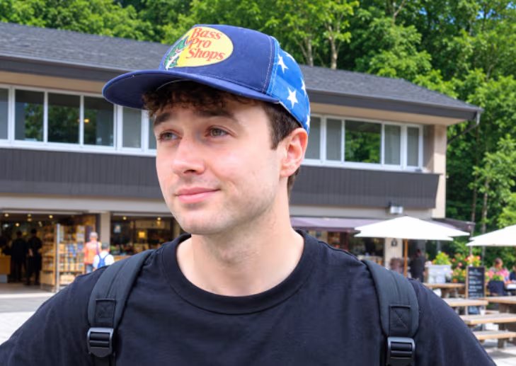 Jacob Coxon posa al aire libre con una gorra azul de béisbol y una mochila frente a un edificio. (@hilbertspaess)