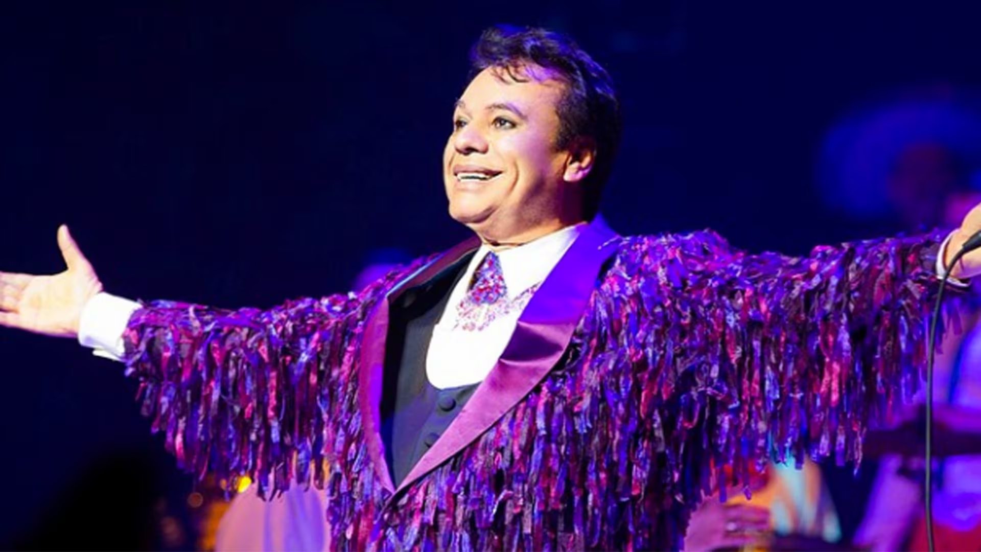 Juan Gabriel sonríe en el escenario con un llamativo atuendo morado.
 